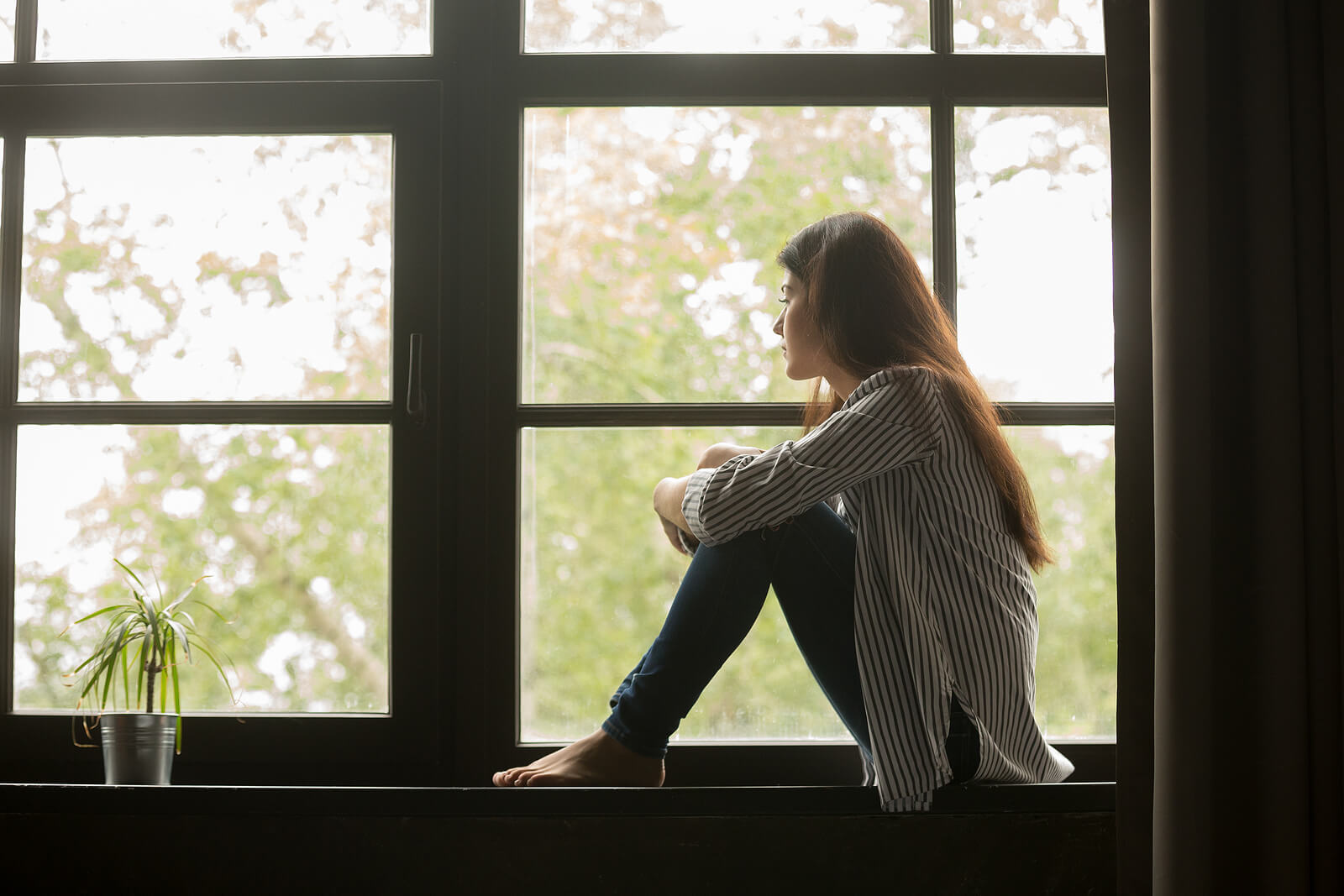 Image of a woman sitting on a window sill and looking out a window holding her knees. Discover how effectively anxiety therapy in Saint Petersburg, FL can help you control your panic attacks and find your triggers.