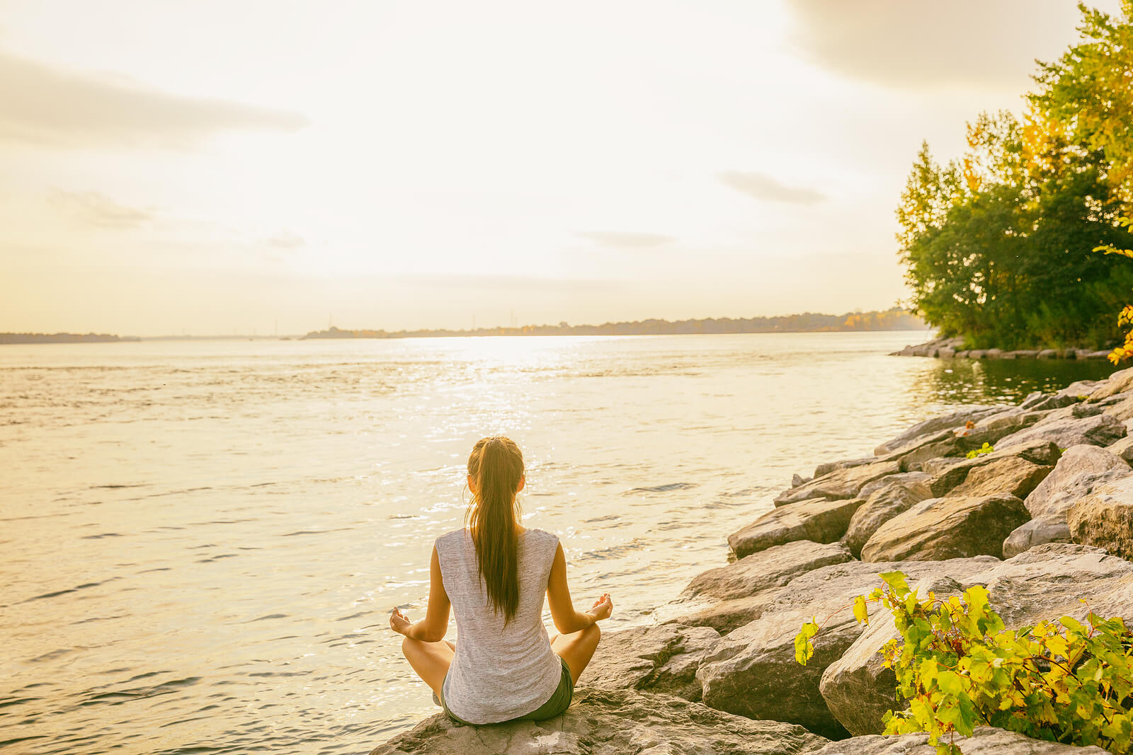 Image of a woman sitting cross legged on a rock in front of a body of water on a sunny day practicing yoga. Learn to uncover your panic attack triggers with the help of anxiety therapy in Saint Petersburg, FL.