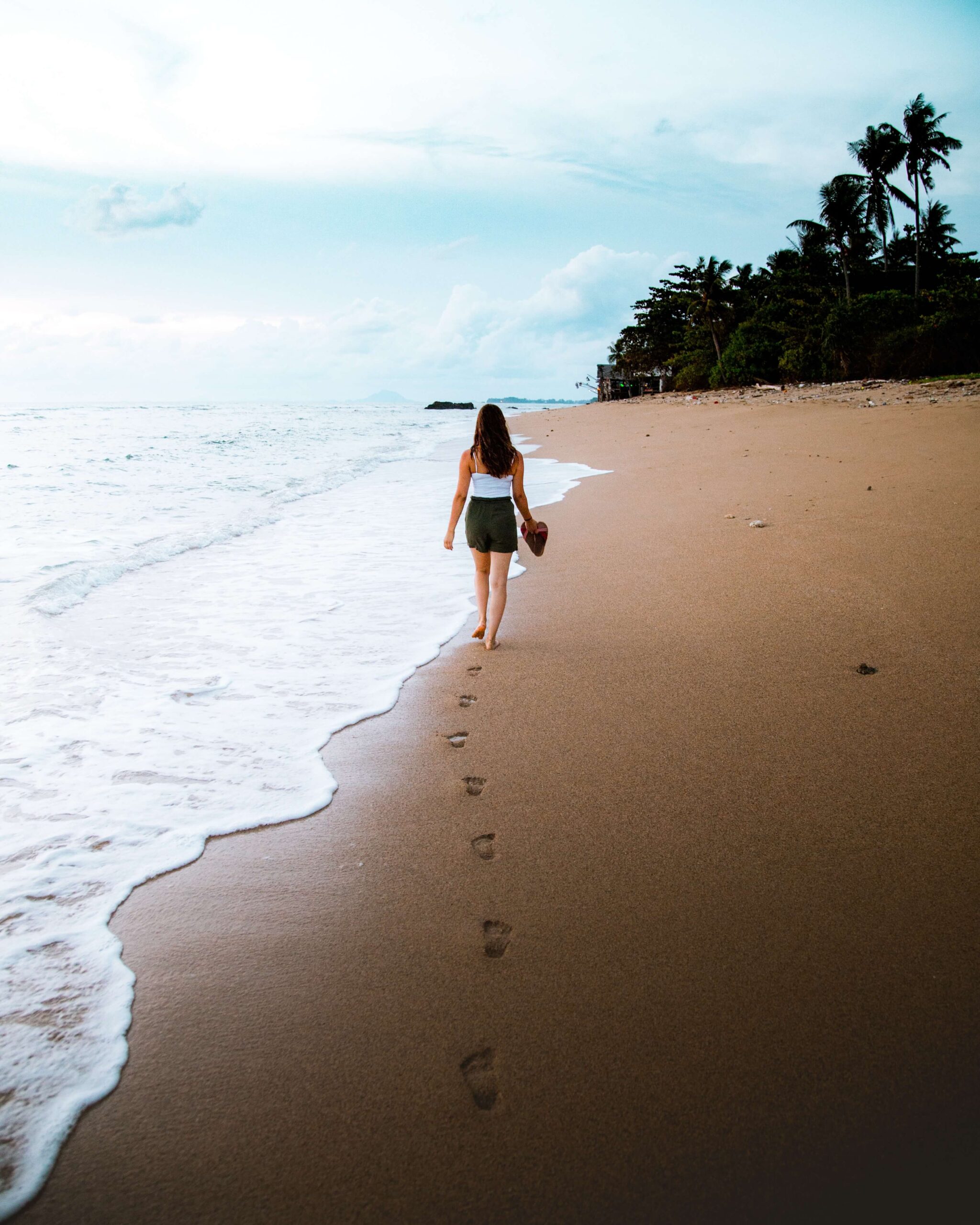 Image of a woman walking down the beach holding her shoes with her footprints in the sand. With the help of a skilled trauma therapist, you can begin managing your PTSD in healthy ways in PTSD treatment in Saint Petersburg, FL.