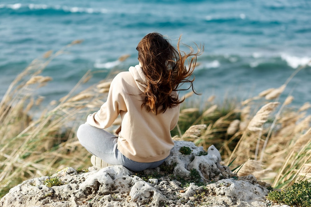 Image of a woman sitting on a rock on a windy day looking out at the ocean. Learn to begin healing and coping with your grief in a healthy way with the help of grief counseling in Saint Petersburg, FL.