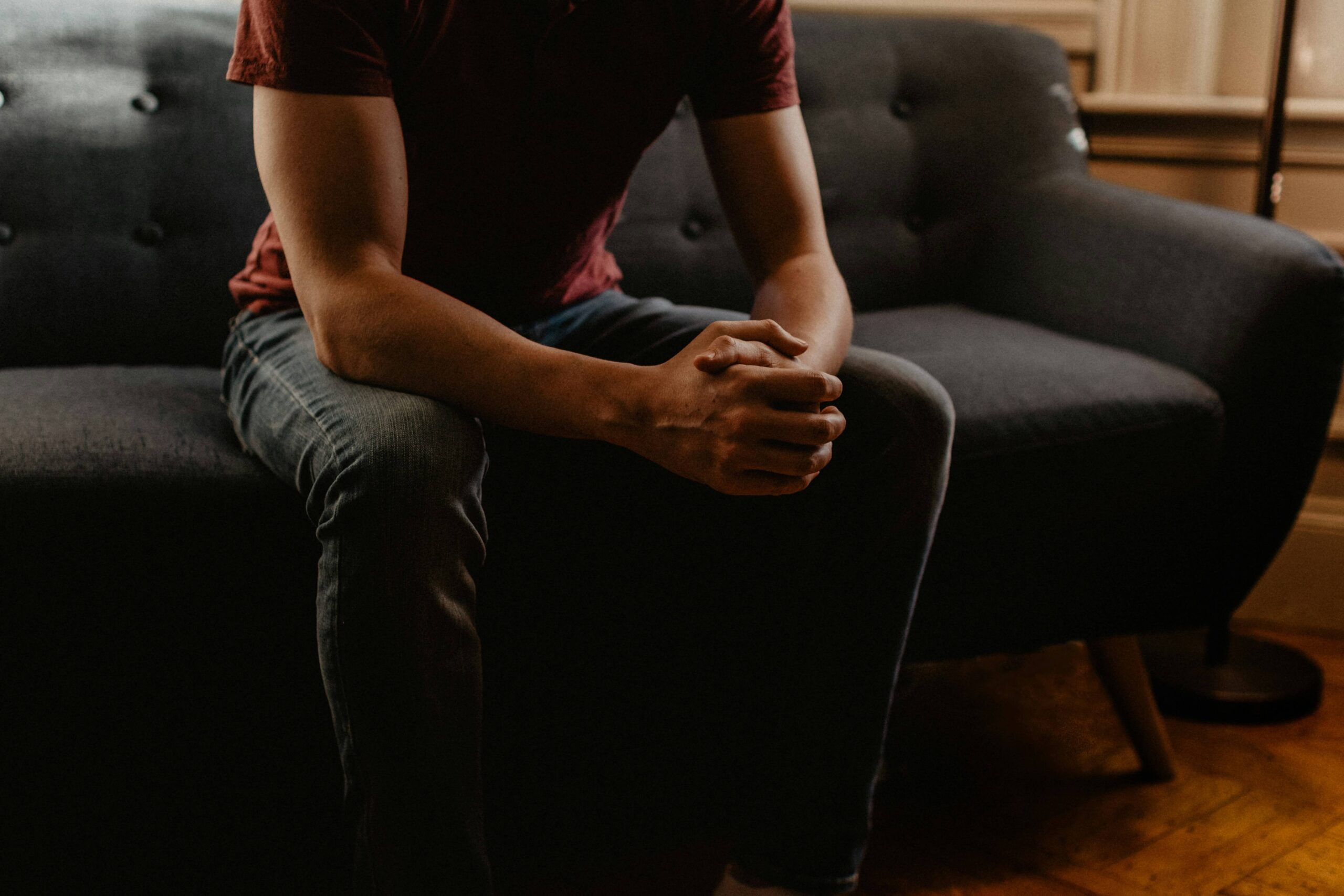 Image of a man sitting on a couch in a dark room, with his hands clasped together. discover how a compassionate grief counselor in Saint Petersburg, FL can help you adjust to the loss of someone.