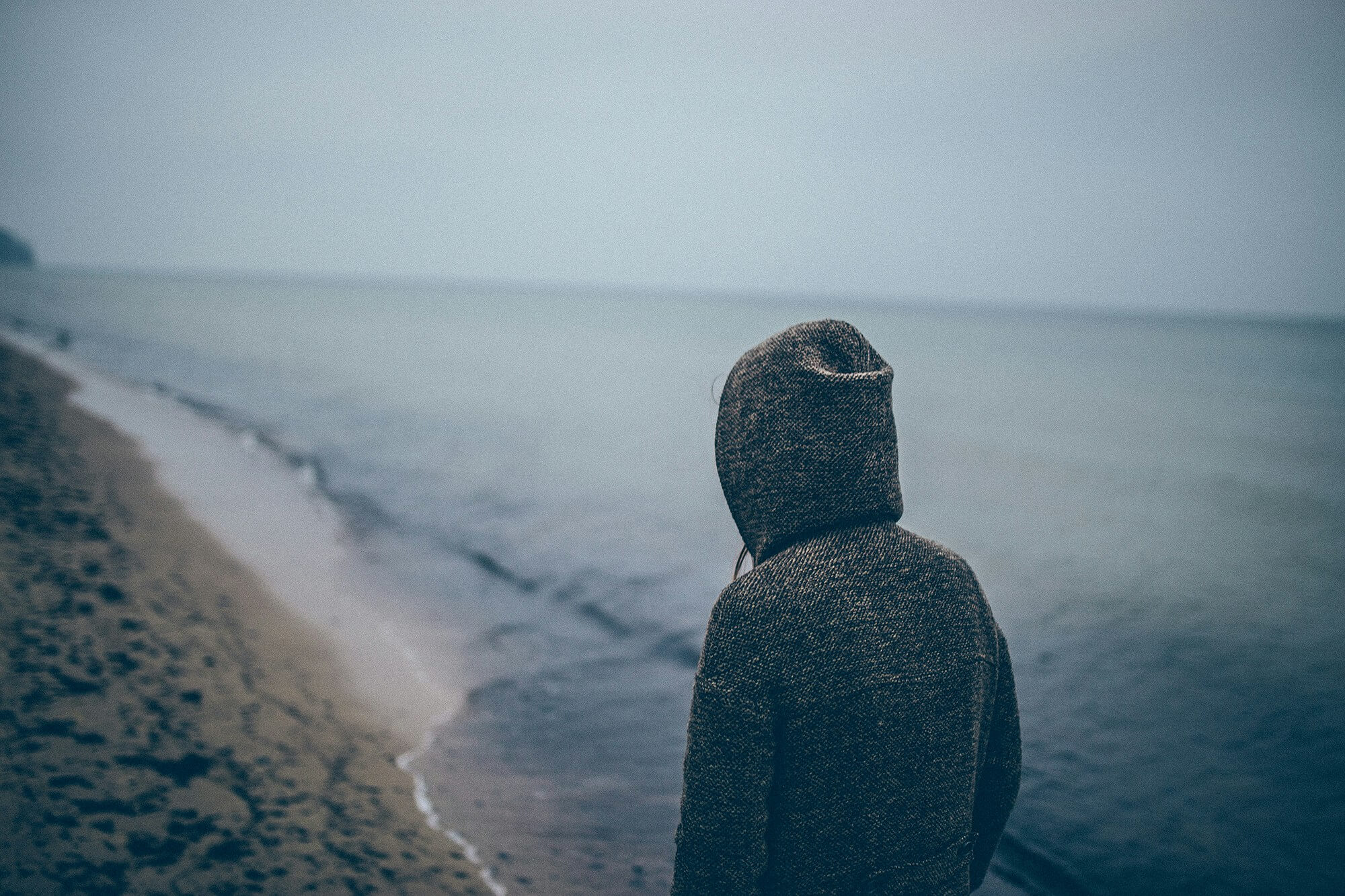 Image of a man standing by the ocean with his hood up on a dark cloudy day. if you struggle to effectively cope with your grief, learn how grief counseling and Saint Petersburg, FL can help provide support.