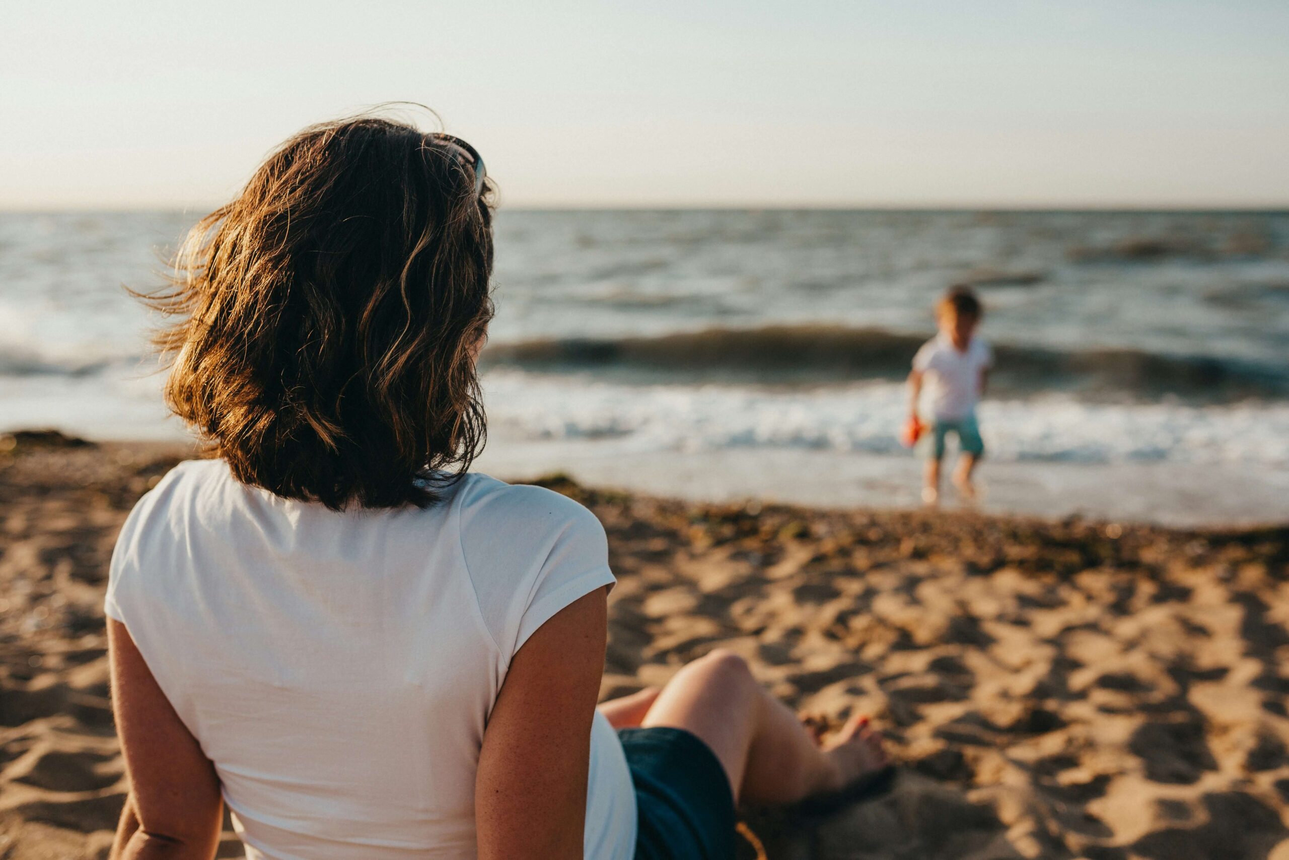 Image of a calm woman sitting on the beach looking at her child playing by the ocean while the sun sets. if you are learning to cope with the loss of a loved, one, discover how grief counseling in Saint. Petersburg, FL can help provide support.